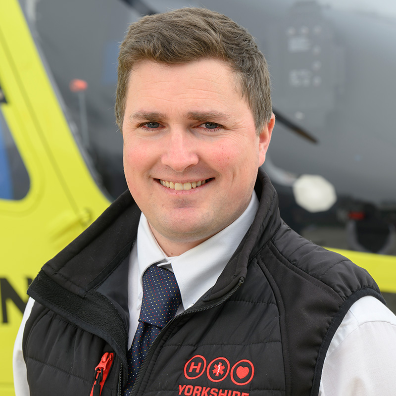 A man with short brown hair, wearing a black gilet over a white shirt, and a blue tie, standing in front of a yellow helicopter.