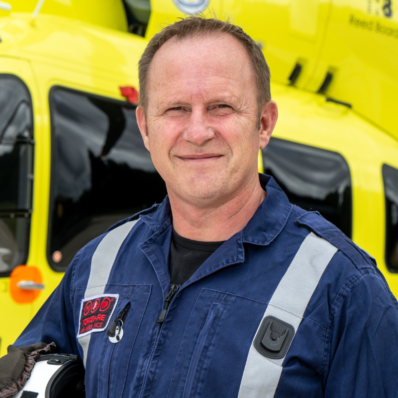 Head and shoulders photo of a man with short brown hair, wearing a blue flight suit, standing in front of a yellow helicopter.