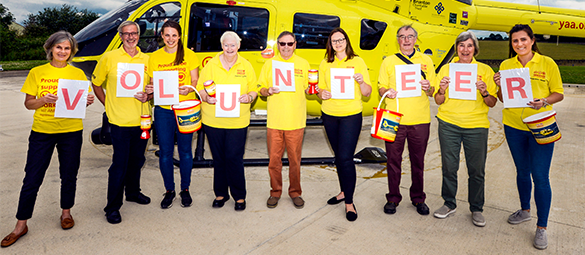 Line of people wearing yellow t-shirts holding up pieces of paper with letters that spell out the word Volunteer, in front of a yellow Yorkshire Air Ambulance helicopter