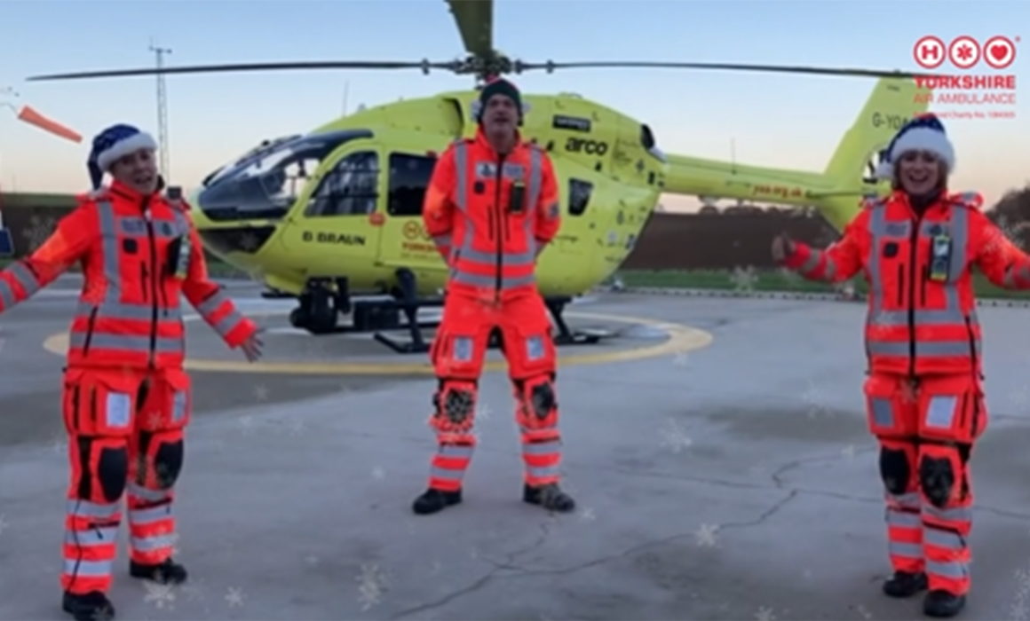 Three members of the Yorkshire Air Ambulance crew stood in front of the yellow helicopter, wearing Christmas hats. The Yorkshire Air Ambulance logo is in the top right hand corner.