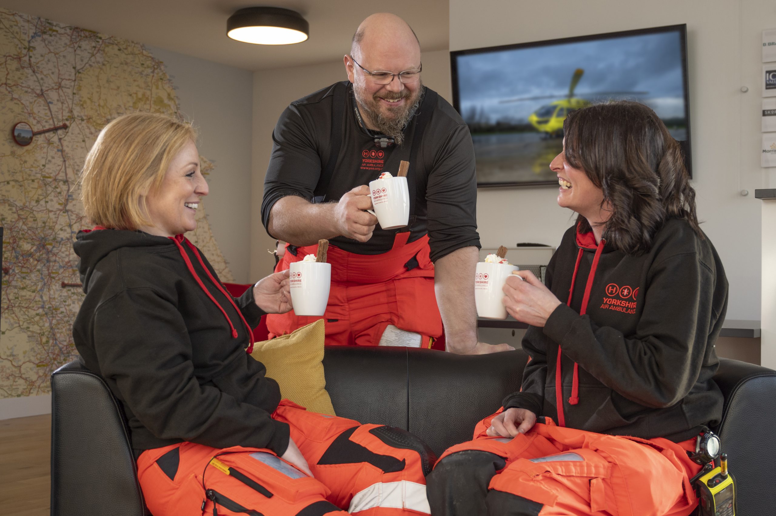 Two members of the Yorkshire Air Ambulance crew are sat on a black couch and one member of the crew is stood behind the couch. They are all holding a cup of hot chocolate and they are all laughing. In the background there is a screen on the wall displaying a picture of the yellow Yorkshire Air Ambulance helicopter.