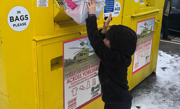 A child wearing a black fleece hoodie is reaching to place a bag into a yellow recycling bank. The recycling bank has a graphic on the front with a yellow Yorkshire Air Ambulance helicopter on it and the words HELP TO SAVE LIVES ACROSS YORKSHIRE