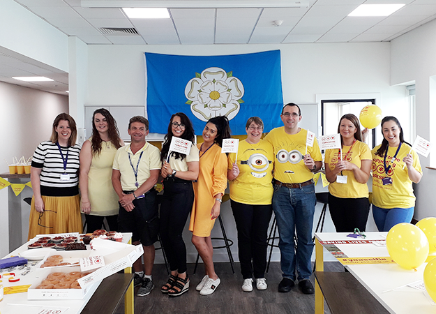 A group of people, wearing mainly yellow clothing, are stood in front of a Yorkshire rose flag. Some are waving small Yorkshire Air Ambulance flags and in front of them is a table with lots of sweet treats on it. To the far right of the image are some yellow balloons.