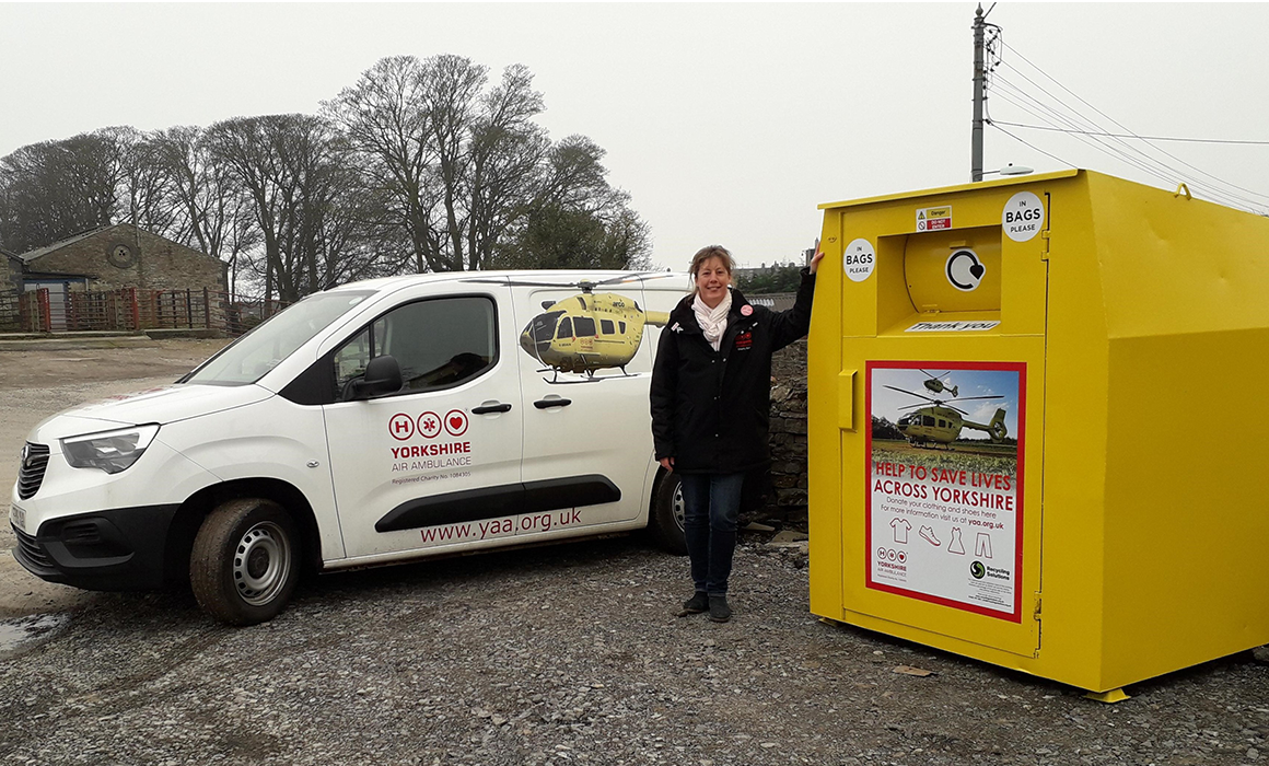 A Yorkshire Air Ambulance fundraiser stands with a yellow Yorkshire Air Ambulance recycling bank, in front of a white van with a picture of a yellow Yorkshire Air Ambulance helicopter on it and the Yorkshire Air Ambulance logo