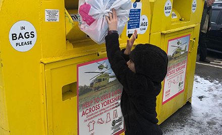 A child placing a bag of recycling in to a yellow Yorkshire Air Ambulance recycling bank