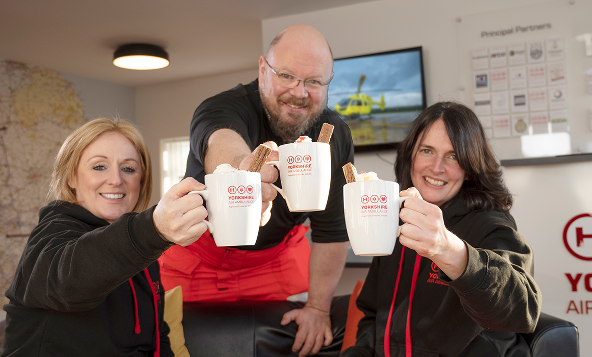 3 members of Yorkshire Air Ambulance crew holding out cups of hot chocolate in Yorkshire Air Ambulance cups with cream on top and a chocolate flake. Two of them are sat down and one is stood behind the couch.