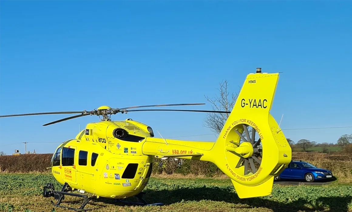 The Yorkshire Air Ambulance yellow helicopter G-YAAC is on a field next to a lane, with a blue car parked in the background, behind the tail of the aircraft. There are some hedges, trees and fields in the background and a power line