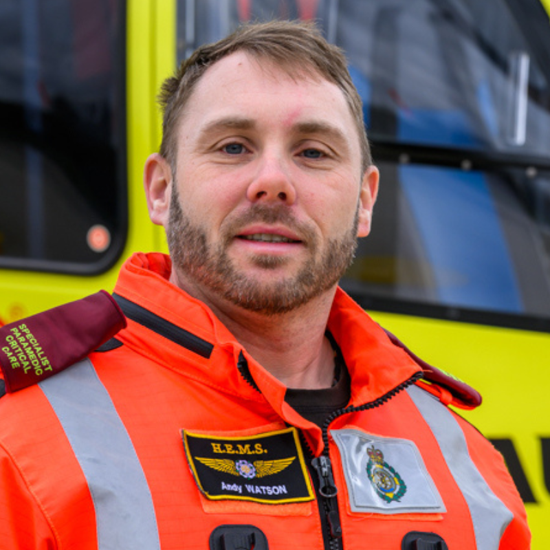 A man with short brown hair wearing an orange flight suit is stood in front of a yellow helicopter.
