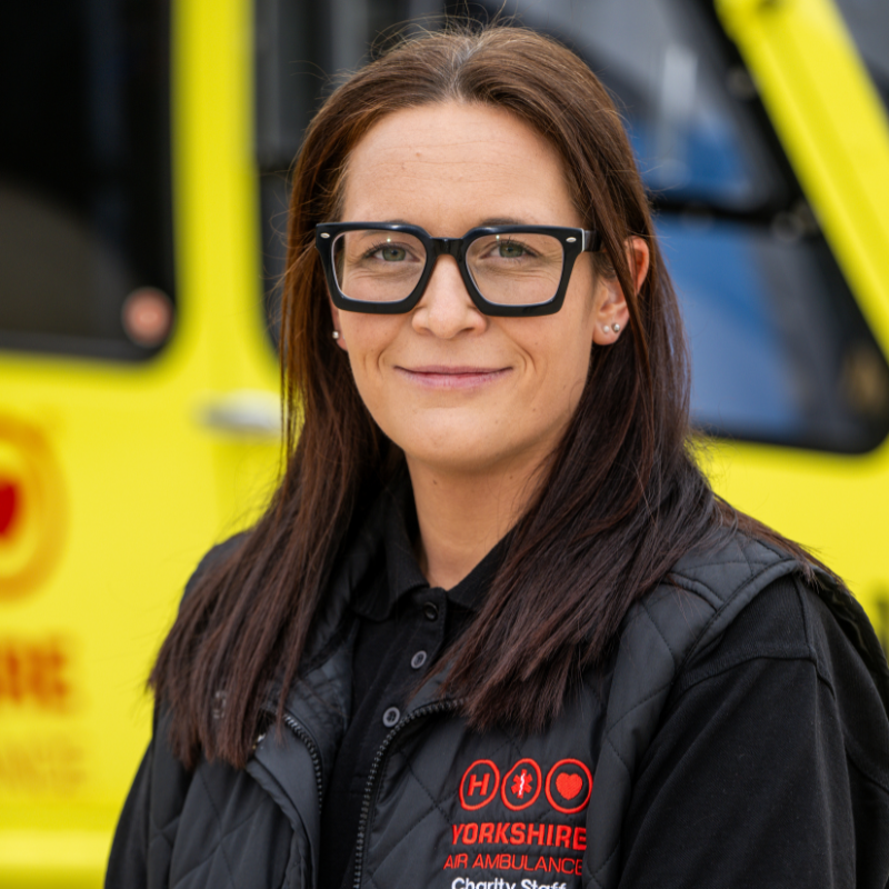 A woman with long straight brown hair, wearing black glasses and a black top with a red logo, is stood in front of a yellow helicopter.
