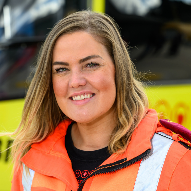 A lady with long brown hair, wearing an orange flight suit is standing in front of a yellow helicopter.