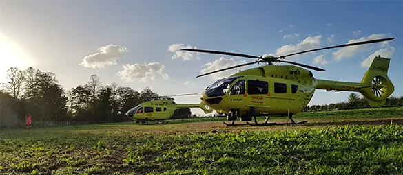 Photo of two yellow Yorkshire Air Ambulance helicopters in a field with the sun shining down
