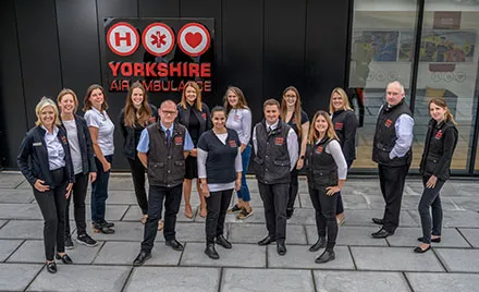 Photo of members of the Yorkshire Air Ambulance fundraising team stood in front of the airbase reception and the Yorkshire Air Ambulance logo