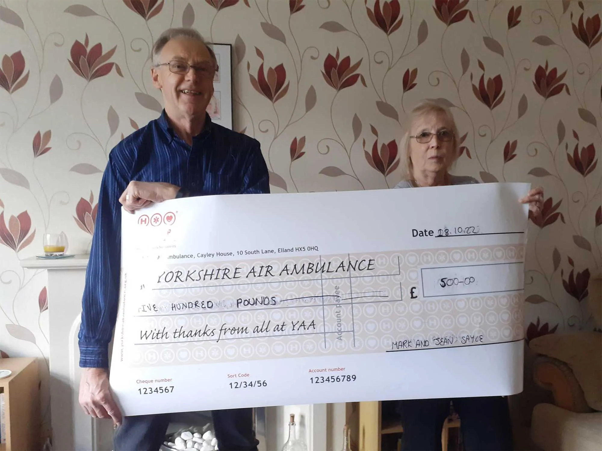 Mark and Jean Sayle are holding a giant cheque. The cheque is made payable to Yorkshire Air Ambulance and is for five hundred pounds. They are stood in front of a wall covered with flowered wall paper.