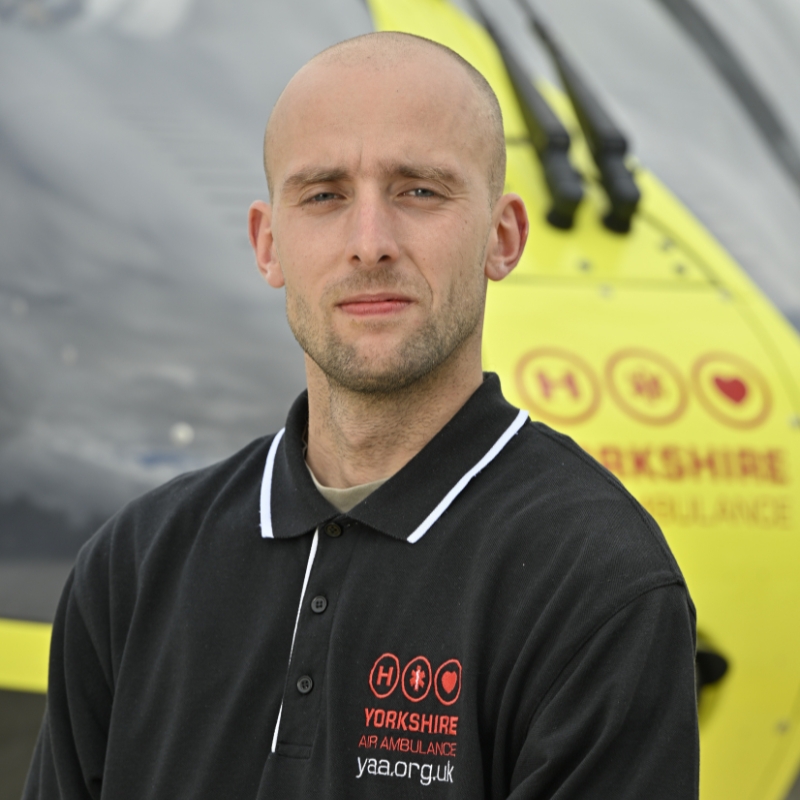 Yorkshire Air Ambulance Technical Crew Member Alex Clark, wearing a black polo shirt, with a white band around the collar, and red Yorkshire Air Ambulance logo standing in front of the yellow Yorkshire Air Ambulance helicopter