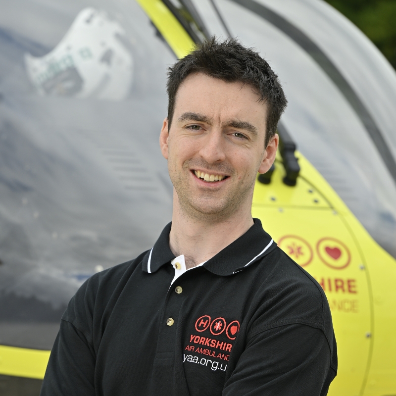 Yorkshire Air Ambulance Technical Crew Member Ross Fairbairn, wearing a black polo shirt, with a white band around the collar, and red Yorkshire Air Ambulance logo standing in front of the yellow Yorkshire Air Ambulance helicopter