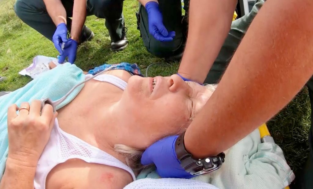 Photo shows a lady, Stella Collins, laying on a stretcher in a field, whilst someone is holding her head still. They are wearing purple gloves and there are two people wearing purple gloves knelt down in the backgroun.