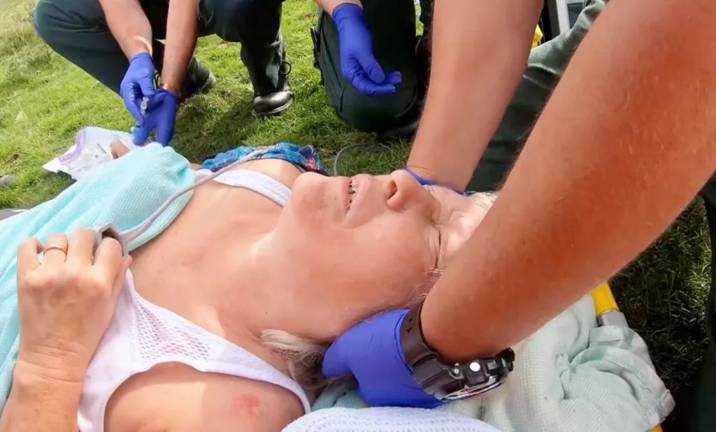 Photo shows a lady, Stella Collins, laying on a stretcher in a field, whilst someone is holding her head still. They are wearing purple gloves and there are two people wearing purple gloves knelt down in the backgroun.
