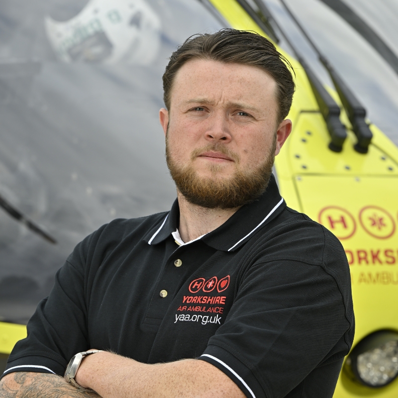 Yorkshire Air Ambulance Technical Crew Member Tom Edge, wearing a black polo shirt, with a white band around the collar, and red Yorkshire Air Ambulance logo standing in front of the yellow Yorkshire Air Ambulance helicopter