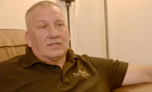 Photo of Robin Brown. The man is sat in a cream coloured arm chair or sofa and is wearing a brown polo shirt with a yellow logo on it. 