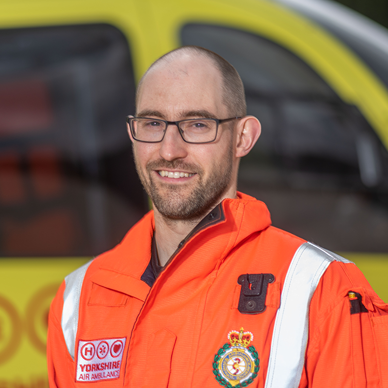 Photo of Dr David Driver wearing an orange flight suit carrying both the Yorkshire Air Ambulance and Yorkshire Ambulance Service logos, stood in front of one of the yellow Yorkshire Air Ambulance service helicopters. Image shows head and shoulders of Dr David only.
