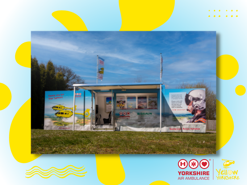 One of the Yorkshire Air Ambulance Promotional vehicles, on grass, with a tree in the background and blue sky with white clouds. The vehicle has two sides out, one varrying an image of two yellow helicopters and the other a side image of a pilot in a fligh helmet with visor and mic. The door to the vehicle is open and there is a canapy out. There are two flags above the vehicle, you can see that the left hand one has an image of yellow helicopters on and the Yorkshire Air Ambulance logo. The image is on a graphic background, which has a blue to white fade and a yellow paint splodge sitting behind the image. There are also some yellow wave and dot graphics and the red Yorkshire Air Ambulance logo with yellow Yellow Yorkshire logo along side it. 