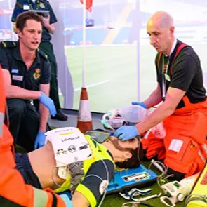Two meant are knelt next to a mannequin. The men are wearing paramedic uniforms and blue gloves. One is holding an air mask on to the mannequin's face. There is another person in the background, their head is not visible. They are also wearing a green paramedic uniform. There appears to be the scene of a football pitch projected on to the wall in the background. There is another piece of medical equipment across the chest of the mannequin.