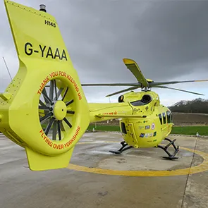 A yellow helicopter stands on a helipad with fields and trees in the background. The image is looking down the body of the helicopter from the tail. The letters 'G-YAAA' are written in black on the tail. The aircraft has 5 rotor blades.