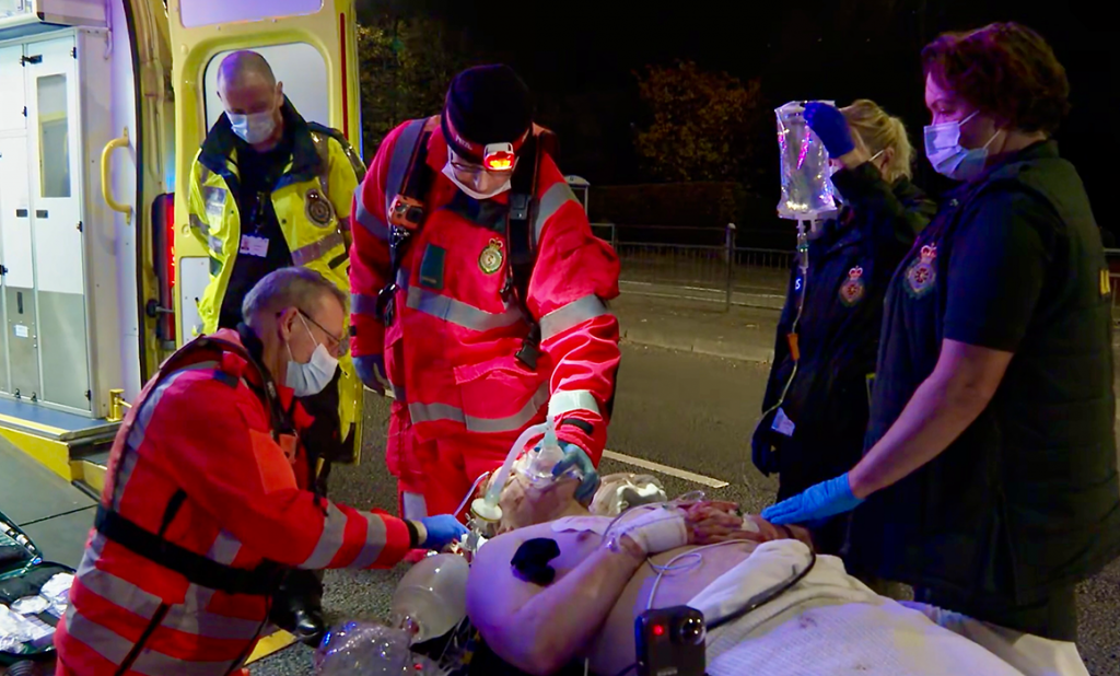 Medical staff are treating a man who is lying on a stretcher. They are in a road and an ambulance is visible in the background. 