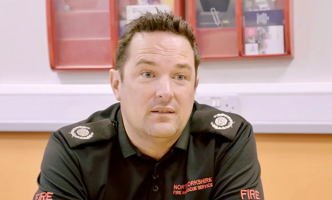 Head and shoulders view of a gentleman with short dark hair. He is wearing a black shirt with the word 'FIRE' on both sleeves in red. On the front right hand chest are the words ' NORTH YORKSIRE FIRE AND RESCUE SERVICE' in red. His shirt has epaulettes on both shoulders, each has a white or silver crest on.