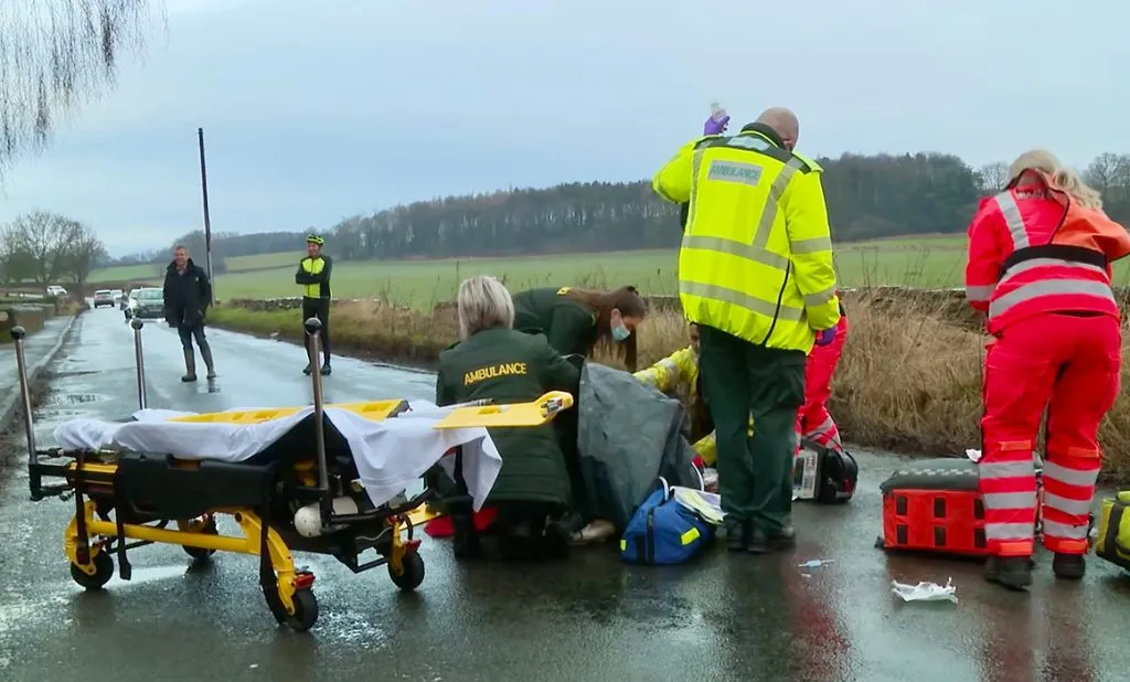 Image shows an accident scene on a wet road. There are fields and trees behind. There are a number of people in the road including six medical personnel. The appear to be tending to someone on the floor. The word 'AMBULANCE' is visible on the back of two of them. Two are wearing dark green jackets. Two are wearing yellow hi visibility jackets, and two are wearing orange jackets and trousers. There are two other people stood in the road further back. One is a gentleman wearing a black jacket and wellies. The other is a gentleman wearing a yellow and black cycling helmet , black and yellow cycling jacket, black leggings and black cycling shoes. There are a number of cars which appear to be waiting behind them. 
