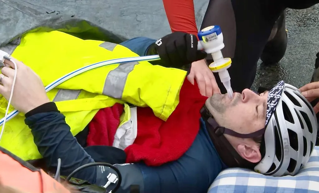 A man wearing a silver cycling helmet is laying down and has a red blanket and a yellow hi visibility jacket over him He has a gas and air pipe in his mouth, which he looks to be holding with his hand, which is wearing a black glove. The hand and knee of another person is visible knelt at the side of him.