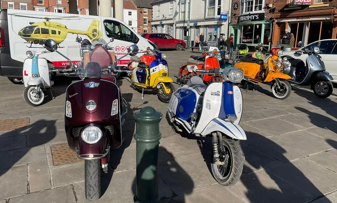 A number of scooters are lined up in a market square, in front of a white van which has a picture of a yellow Yorkshire Air Ambulance helicopter on it