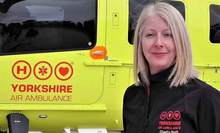 A lady with shoulder length blonde hair, wearing a black coat which carries the red Yorkshire Air Ambulance logo, stands in front of a yellow Yorkshire Air Ambulance helicopter which also carries the red Yorkshire Air Ambulance logo.