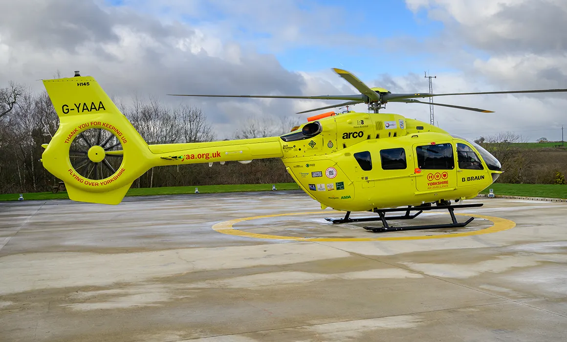 A yellow helictoper sits on a helipad. The image is the side view of the helicopter and it's tail carries the letter 'G-YAAA'. There is a red Yorkshire Air Ambulance logo on the side of the helicopter. There are trees and fields in the background.