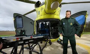 A man with short brown hair and wearing glasses and a green coat, stands in front of a yellow helicotper. He is at the back of the helicopter and the back doors are open. There is a stretcher on a stand and wheels next to him. 