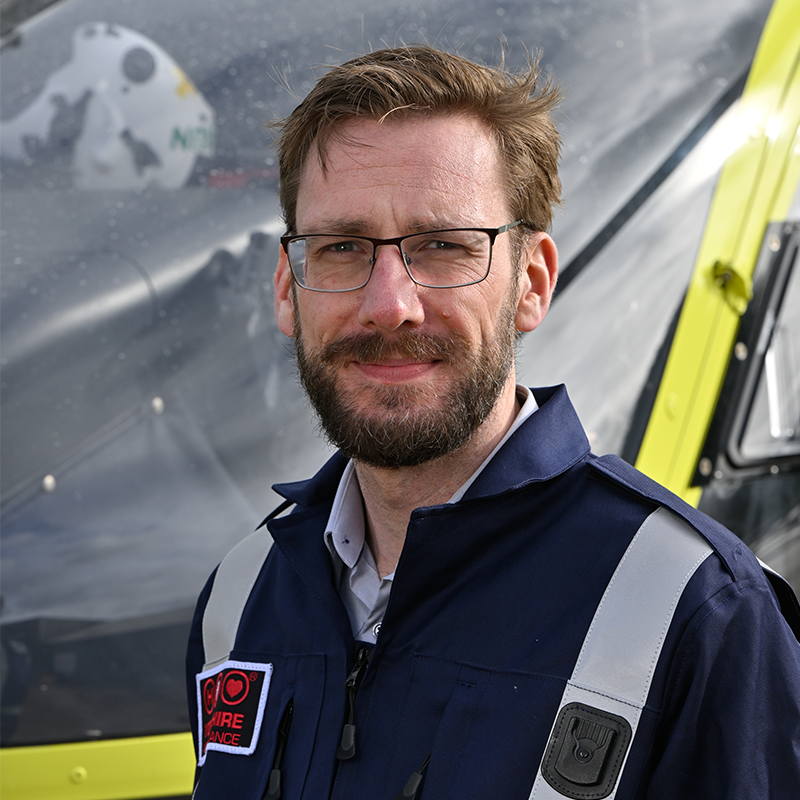 Head and shoulders photo of Yorkshire Air Ambulance Pilot, James Booth. He has short brown hair and is wearing glasses. He has brown facial hair. He is wearing a dark blue flight suit with carries the Yorkshire Air Ambulance logo. One of the yellow helicopters is visible in the background.