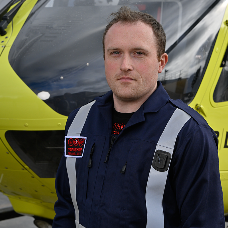 Head and shoulders photo of Yorkshire Air Ambulance Pilot, Josh Adams. He has short brown hair. He is wearing a dark blue flight suit with carries the Yorkshire Air Ambulance logo. One of the yellow helicopters is visible in the background.