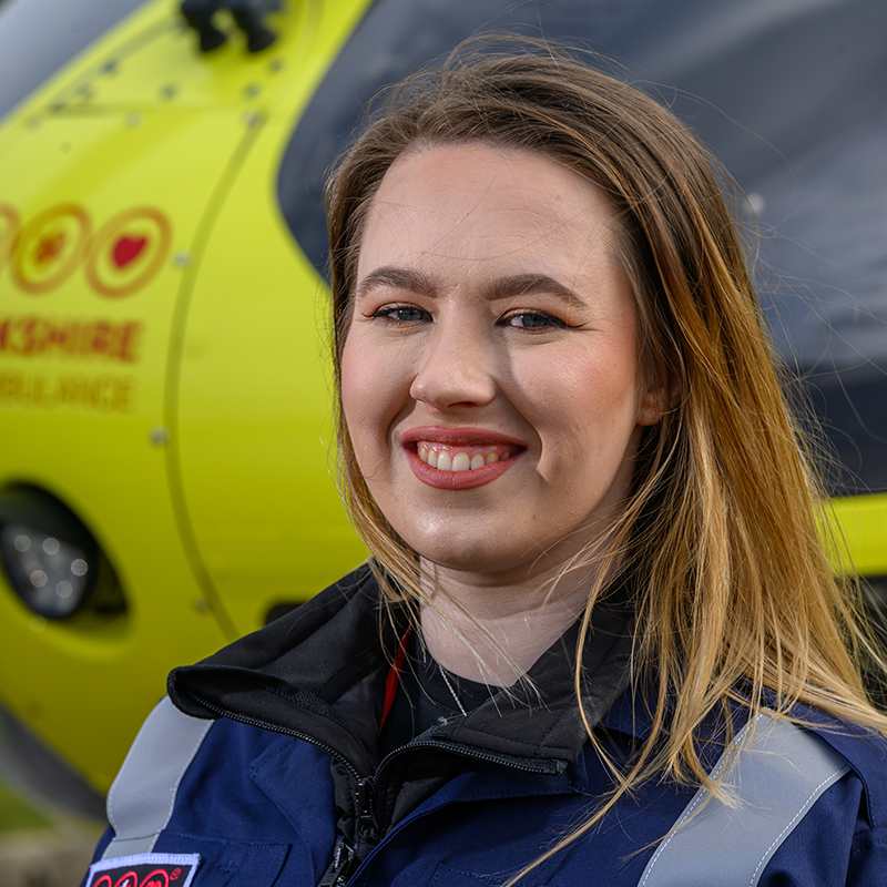 Head and shoulders photo of Yorkshire Air Ambulance Technical Crew Member, Loz Lyles. She has long brown hair and is wearing a dark blue flight suit with carries the Yorkshire Air Ambulance logo. One of the yellow helicopters is visible in the background.