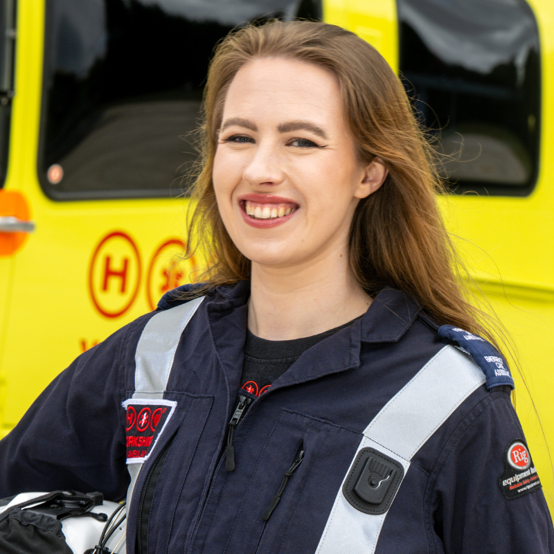 Head and shoulders photo of a woman with long brown hair, wearing a blue flight suit and standing in front of a yellow helicopter.