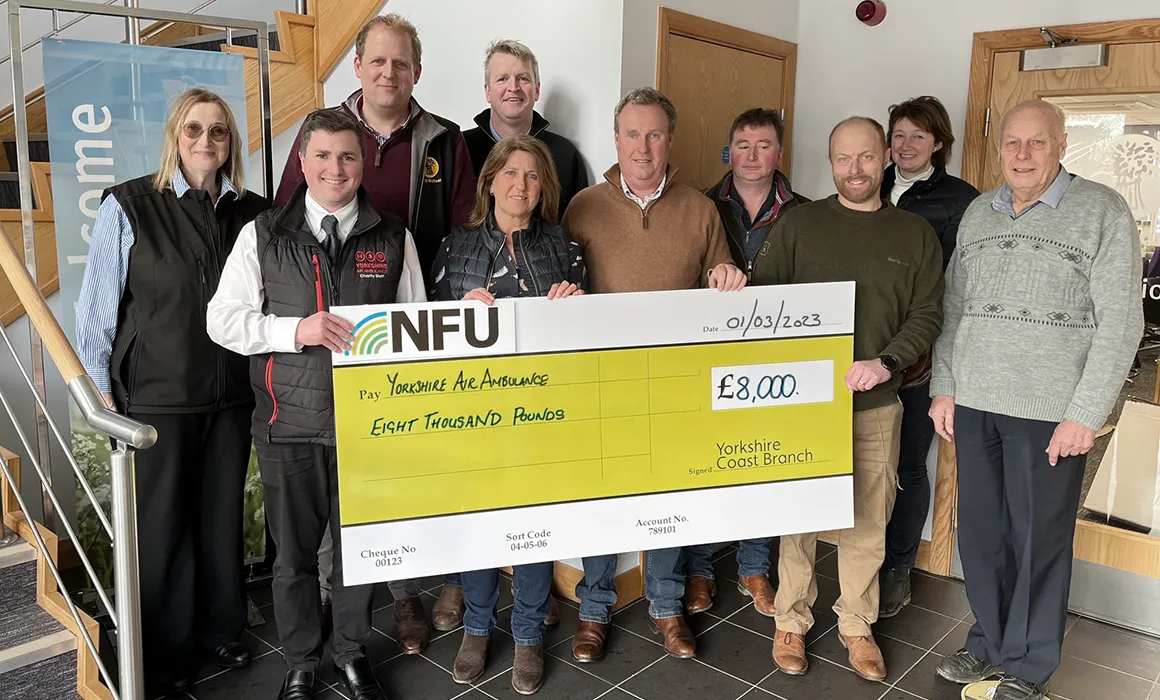 A group of people stood in front of a wooden and glass staircase and a wooden door. They are holding a large yellow and white cheque which carried the NFU logo and is made payable to Yorkshire Air Ambulance with a figure of £8000