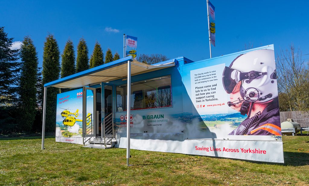 Photo shows one of the Yorkshire Air Ambulance promotional vehicles parked on grass. The sides are out and display photos of the yellow helicopter and a pilot with a helmet on and has some wording, which cannot be made out on the photo. The words Saving Lives Across Yorkshire are along the bottom. There is an awning out from the side of the vehicle. There are open doors with steps and a large window. On top of the vehicle are two flags.