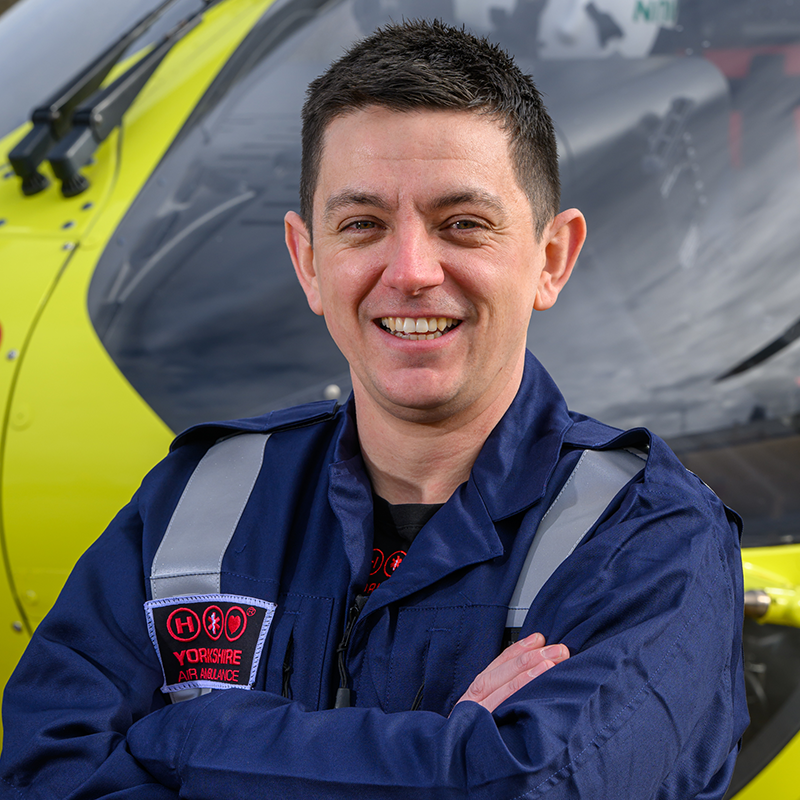 Head and shoulders photo of Yorkshire Air Ambulance Technical Crew Member Stephen Kenworthy. He has short brown hair and is wearing a dark blue flight suit with carries the Yorkshire Air Ambulance logo. One of the yellow helicopters is visible in the background.