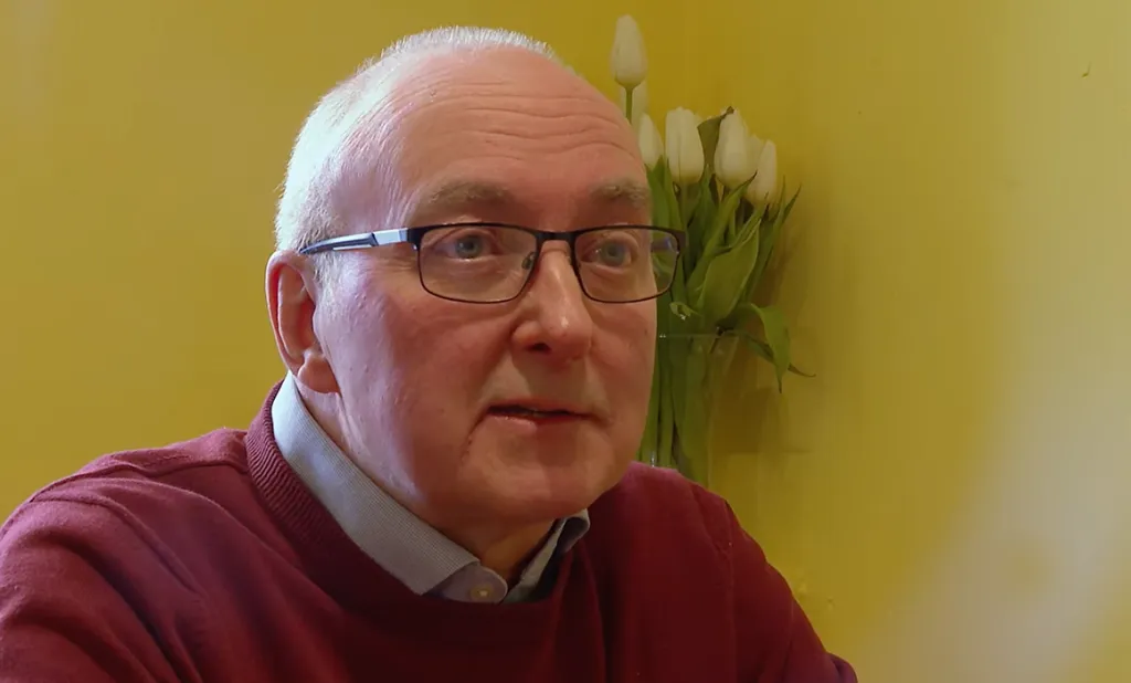 Head and shoulders images of Stephen Curran. He is wearing a burgundy jumper with a shirt collar visible. He has short fair coloured hair and is wearing glasses. He is sitting in front of a cream coloured wall and there is a vase in the background containing white or cream tulip flowers.