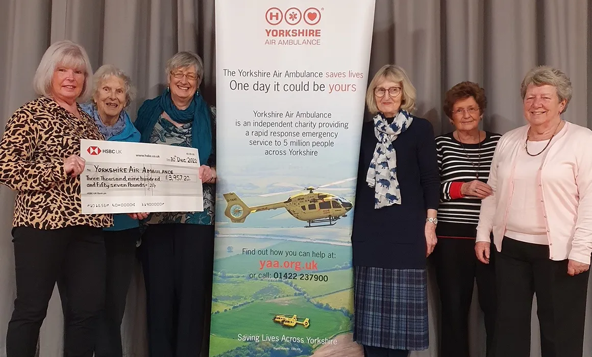 Six members of Thirsk Ladies Club are standing next to a Yorkshire Air Ambulance banner and holding a large cheque for £3957.20 made payable to Yorkshire Air Ambulance. They are standing in front of a big grey curtain.