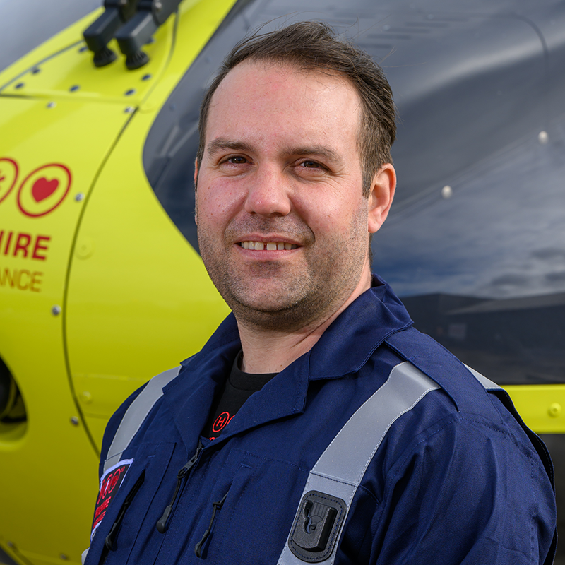 Head and shoulders photo of Yorkshire Air Ambulance Technical Crew Member Will Collinson. He has short brown hair and is wearing a dark blue flight suit with carries the Yorkshire Air Ambulance logo. One of the yellow helicopters is visible in the background.