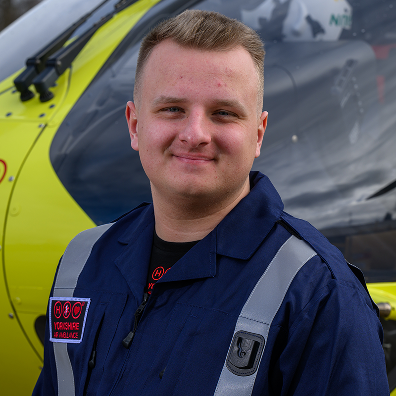 Head and shoulders photo of Yorkshire Air Ambulance Technical Crew Member Will Newton. He has short fair hair and is wearing a dark blue flight suit with carries the Yorkshire Air Ambulance logo. One of the yellow helicopters is visible in the background.
