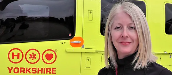A lady with shoulder legnth blonde hair and wearing a black top is standing in front of a yellow aircraft which carries the red Yorkshire Air Ambulance logo. Only part of the aircraft is visible, some of the yellow body and logo and parts of three black windows.
