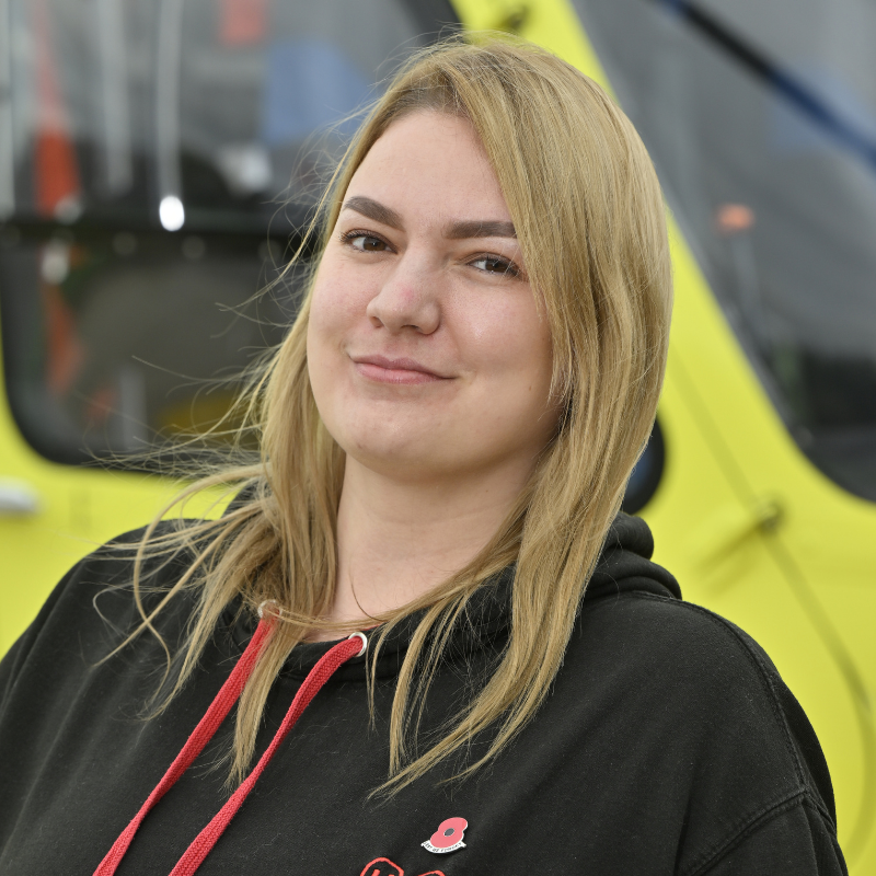 A head and shoulders image of a lady with longe blonde hair, wearing a black hoodie with red trim standing in front of a yellow helicopter.