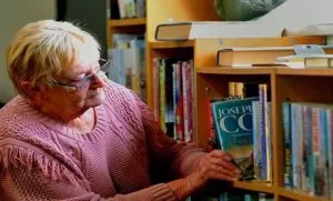 A lady with short light hair, wearing a pink jumper places a book on to a shelf containing other books.