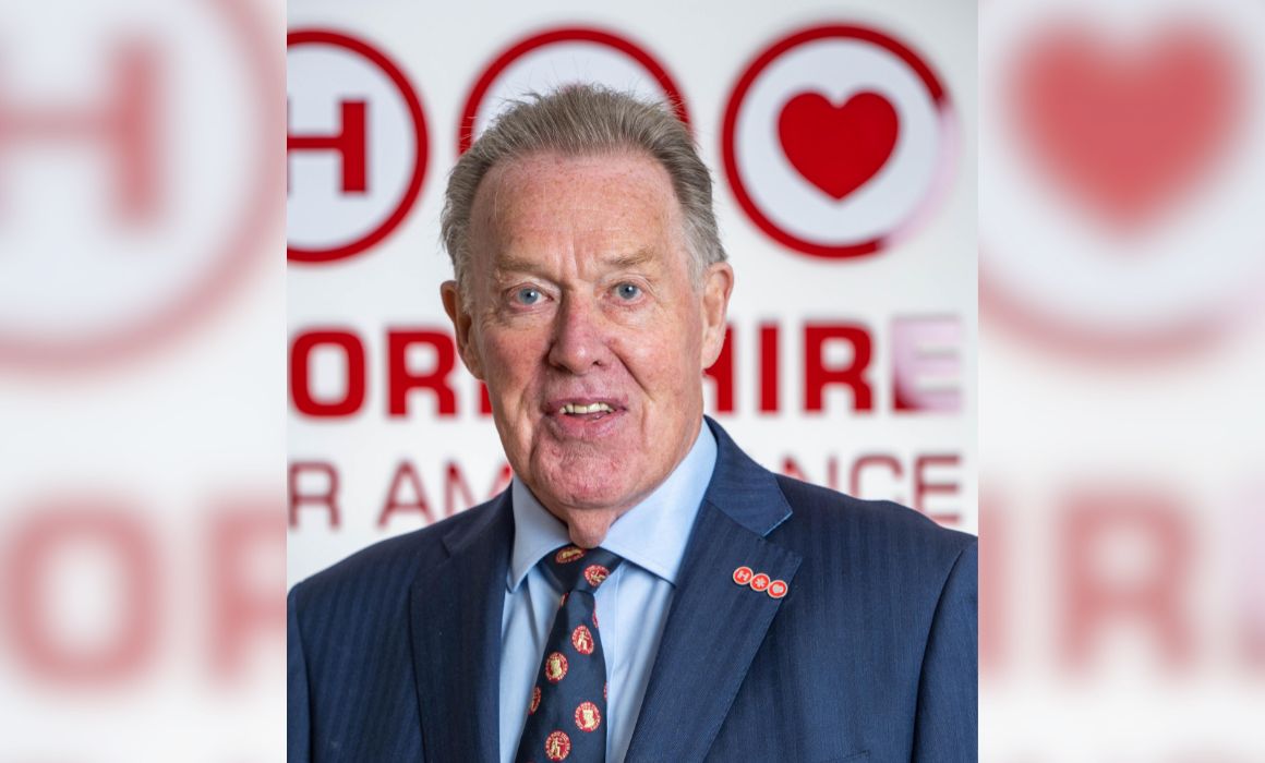 A gentleman wearing a dark blue suit jacket, light blue shirt and a blue tie with a red repeat pattern stands in front of a white background and a large red Yorkshire Air Ambulance logo. The gentleman also has a lapel pin which is the Yorkshire Air Ambulance logo.